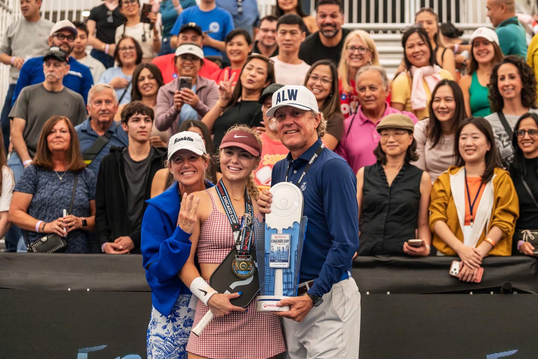 A player wearing a Veolia Chicago Cup gold medal holds her paddle beside the event trophy, posing with two people in front of a crowd of smiling fans in the stands.