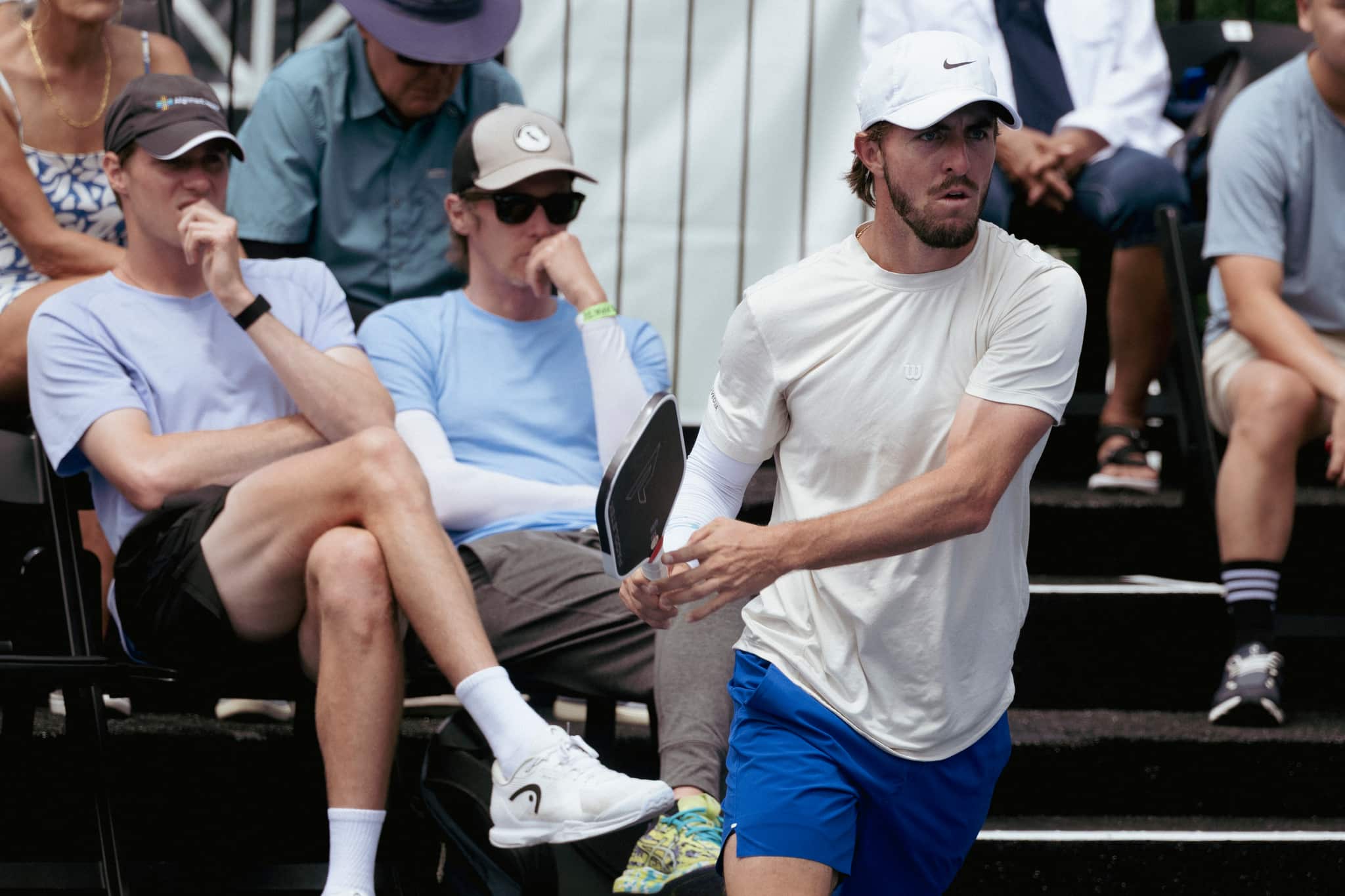 A pro player in a white cap moves to his right with his paddle back, spectators seated in the stands behind him.